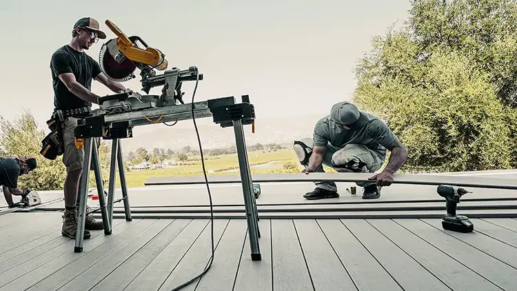 A TimberTech deck contractor cuts a deck board to size while another crouches down to inspect each board