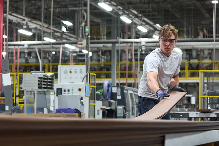 A man wearing safety goggles and gloves works in a factory, handling a decking plank. Bright lights and industrial machines fill the busy workspace.