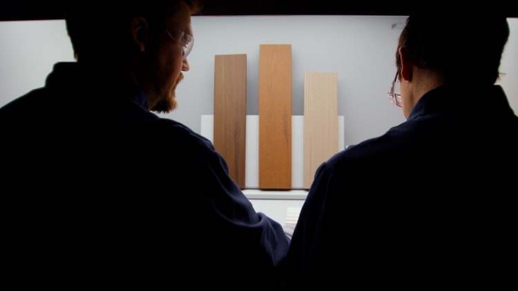 Two people in silhouette examine samples of TimberTech decking against a bright background. Three vertical panels display different wood textures and shades.
