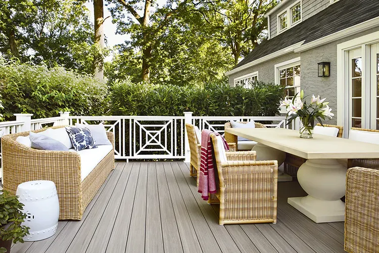 A cozy outdoor patio features a wicker sofa with blue and white pillows, a large beige dining table with wicker chairs, and a vase of flowers. Lush greenery surrounds the space, creating a tranquil atmosphere.
