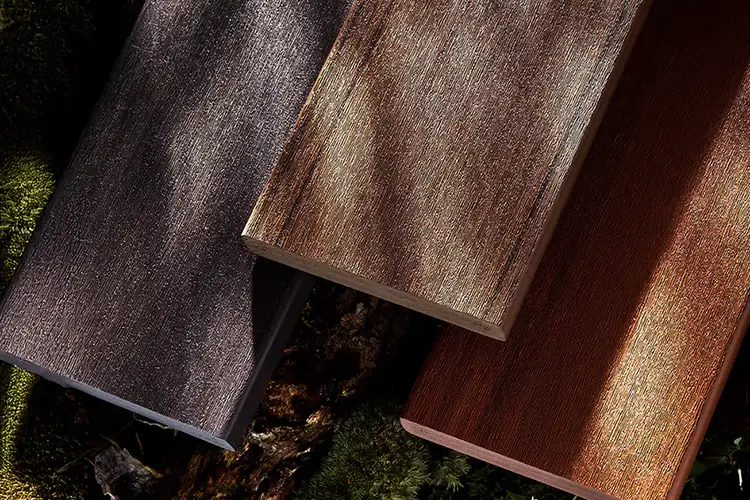 Three rectangular pieces of textured PVC decking in different shades—dark gray, brown, and reddish-brown—placed on a forest floor with moss.