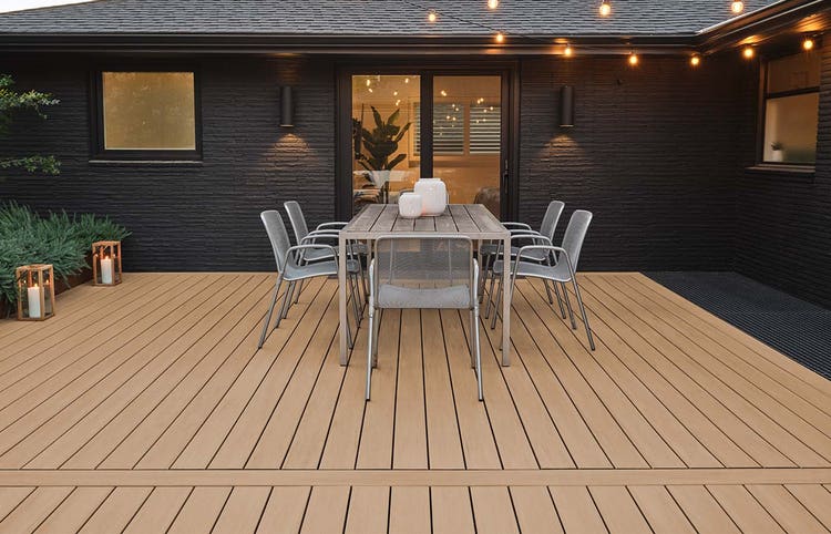 TimberTech Advanced PVC Vintage Collection decking in Weathered Teak creates an evening patio with a modern dining table, black chairs, glowing lanterns, and warm string lights.