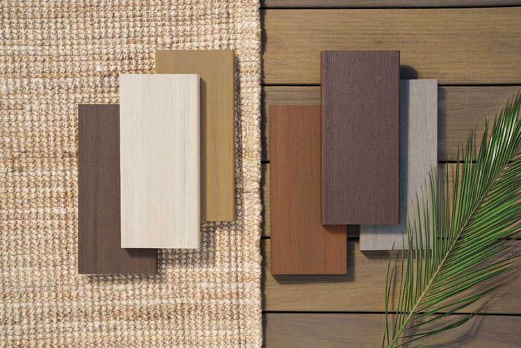 Six TimberTech decking samples in two groups of three display varied wood-grain patterns in brown, tan, beige, and light gray on a woven surface and wood planks beside a green palm leaf.