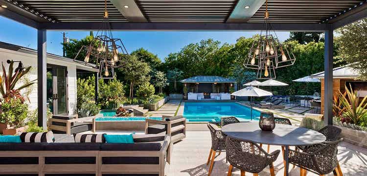 A large pergola sports recessed lights and two contemporary light fixtures hanging above poolside dining and seating areas.