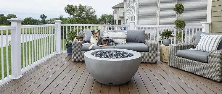 A dog lounges on outdoor furniture in front of a firepit on a deck.