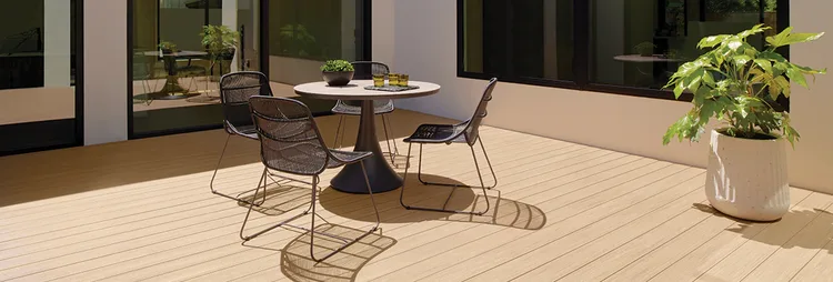 Modern home with floor to ceiling glass doors and large windows has a tan colored deck with a circular table and four chairs around it a few feet from a large potted plant.