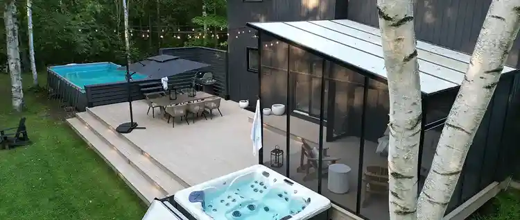 Backside of a modern home with a screened-in outdoor sitting area features a light tan deck with an above ground pool and hot tub