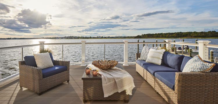 A lakeside deck with glass railing has a matching furniture set including an outdoor sofa, coffee table, and armchair.