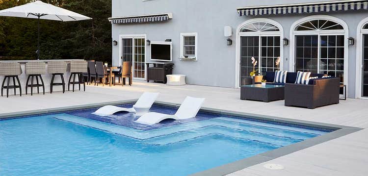 A pool with two submerged lounge chairs shows a deck sectional behind it and adjacent bar and dining area to the left.