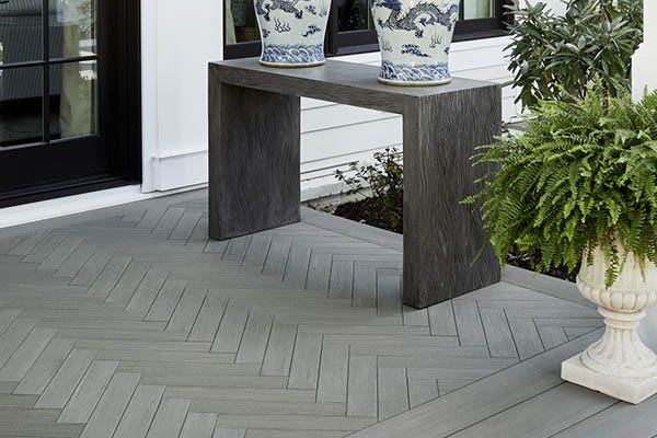 A section of a composite deck just near the entryway to the home features a tight herringbone pattern. A sleek modern table sits on the deck, with two ornate porcelain vases on it. A potted plant in a stone planter adds more texture to the space. The composite deck is made of Coastline Multi-Width Decking from the TimberTech AZEK Vintage Collection.