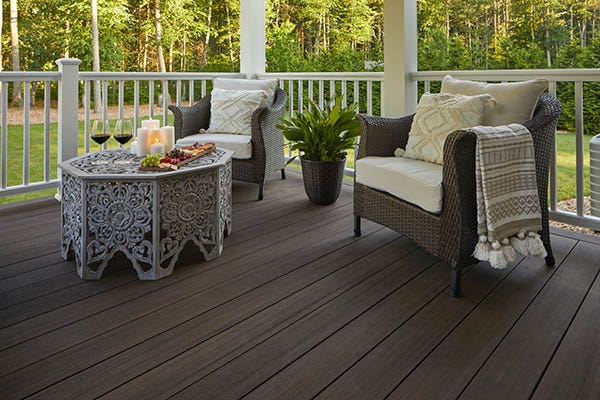 A composite deck is protected under an overhead structure. Two elegant wicker chairs with curved armrests feature dark frames that match the deeply saturated brown decking and white cushions that match the white composite railing. White throw pillows with textured patterns lend dimension to the space. A patterned throw blanket is draped over an armrest. The composite deck is made of American Walnut boards from the TimberTech AZEK Landmark Collection. The railing is the Classic Composite Series Premier Rail in White.