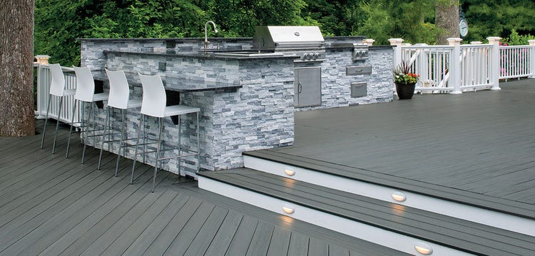 An l-shaped stone kitchen outdoors features a grill and sink, as well as a counter with seating separating the cooking and living spaces.