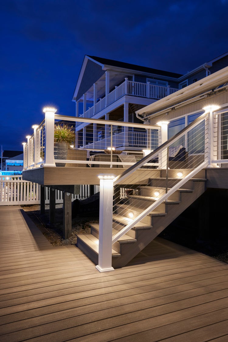 A composite deck is built along a dock. Stairs lead from the deck to the dock. A white composite railing runs along the perimeter of the deck and stairs, and features a stainless steel cable railing infill. Post cap lights illuminate the perimeter and riser lights increase visibility on the stairs. The composite deck is made of Coastline boards from the TimberTech AZEK Vintage Collection.