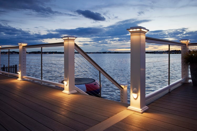A composite deck is built just off the water of a calm bay on the Atlantic coast. A white composite railing with a deck board top rail and stainless steel cable infill runs around the perimeter of the deck and down the stairs to the lower level The railing features lighted island caps and accent lights at the bottom of the boards for ambient illumination. The sky’s light blue and yellow colors suggest it is just after sunset. The gray composite decking is Coastline from the TimberTech AZEK Vintage Collection. The railing is the Classic Composite Series Drink Rail in White with a Coastline deck board top rail.