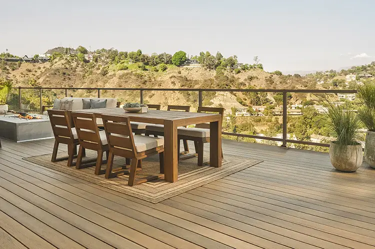 A TimberTech Composite Deck from the Legacy Collection in the color Tigerwood. The light brown, multi-tone deck also features Impression Rail Express railing with Glass Infill to show off the view of the Desert. A wooden dining set sits upon the deck.