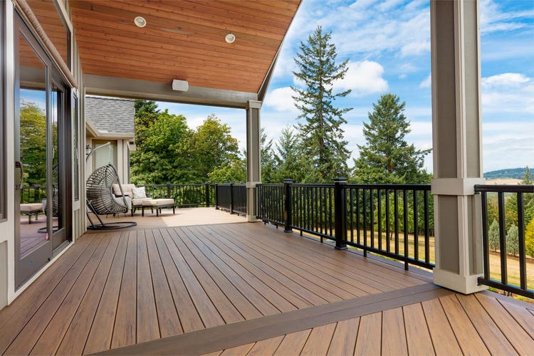 A second-story composite deck runs the length of a home overlooking a hill. An egg chair and plush outdoor chair sit on one end of the deck. Part of the deck is covered by a roof extension, offering coverage from inclement weather. Evergreen trees rise up next to the deck, lending their color and texture to the space.. The composite deck is made of Tigerwood boards with Mocha accents, both from the TimberTech PRO Legacy Collection.