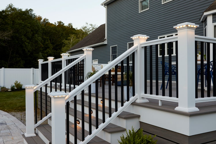 A composite deck features gray composite decking and a white composite railing with black composite balusters. The gray decking is also used as deck skirting in a horizontal application. White PVC trim used as stair risers and fascia creates crisp, clean lines for the deck perimeter. Built-in lighting in the railing posts and stair risers give the scene a romantic glow. The composite deck is made of Coastline boards from the TimberTech AZEK Vintage Collection. The railing is the Classic Composite Series Premier Rail in White.