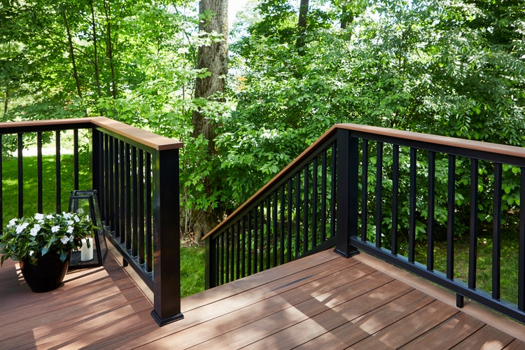 A composite deck is built in a forested area. Sunlight filters through the trees above, leaving dappled light on the deck. A black composite railing runs along the perimeter of the deck and down the stairs. The railing features a deck board top rail that matches the rich brown composite decking of the deck surface. The deck is made of Mahogany boards from the TimberTech AZEK Vintage Collection. The railing is the Classic Composite Series Drink Rail in Black.