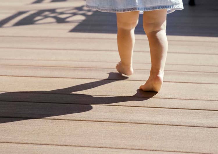 The legs and feet of a toddler in a denim dress are shown walking barefoot on a deck
