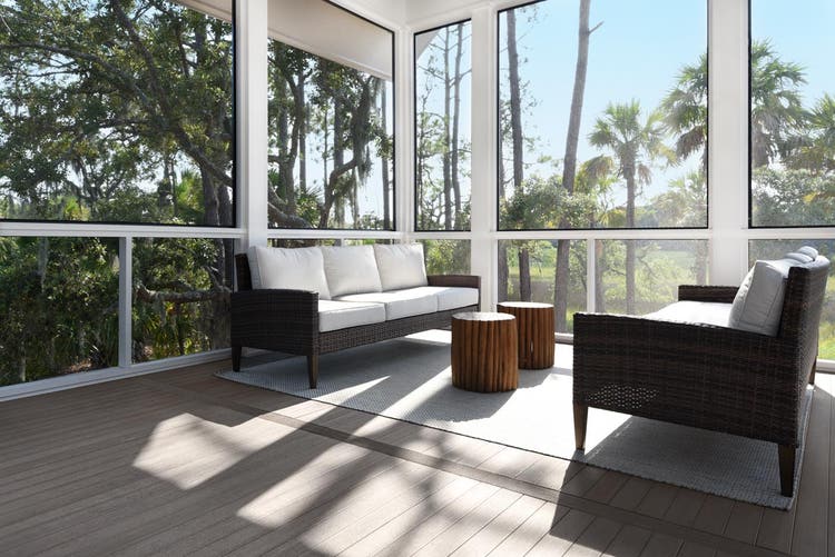 A sunlit, screened porch with large windows, featuring two wicker sofas with white cushions facing each other over a light carpet. The gray Coastline decking is elegantly done in TimberTech Porch Collection decking. In the center, there are two round warm brown wooden tables.