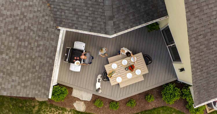 An aerial view of Deb and Rob's deck area that includes a grill and a tan table with plates and benches. Their white and brown dog circles the table as Rob places some table decor on the table.