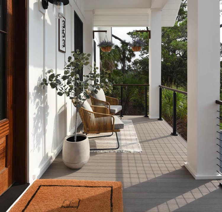 A front porch featuring a doormat, two wicker chairs with cushions, a potted plant, hanging plants, and a house number