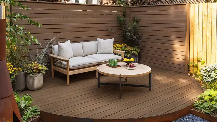 The shaded backyard deck with a wood-framed patio couch with tan pillows and green plants on a medium brown English Walnut PVC deck at the Real Simple NY brownstone home.