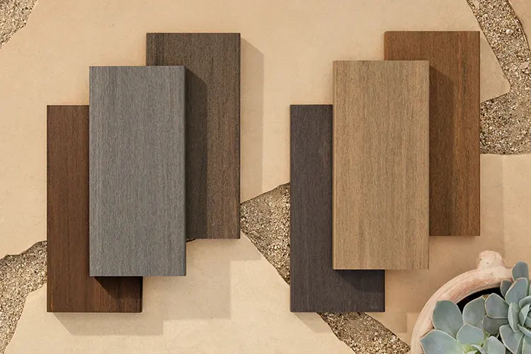 TimberTech deck board samples in brown, gray, and tan shades are arranged on a textured, cracked sandy surface with small stones and succulents to the right.