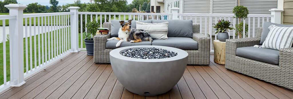 A fire pit is centered on a deck and surrounded by outdoor furniture and decor with a white railing perimeter.