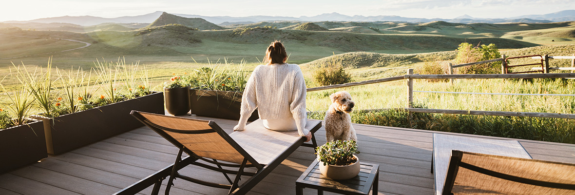 A woman and her dog sit on a deck at golden hour and look out towards rolling hills.