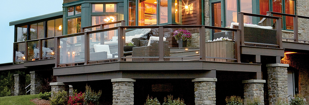 An elevated deck with glass railings and stone pillars wraps around the back of a home with landscaping filling the under deck space