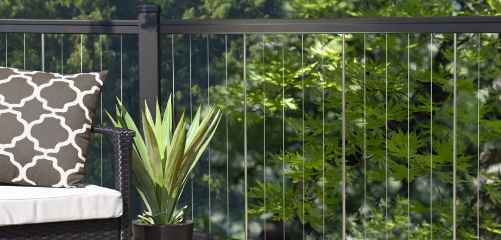 A close-up of a black rail with vertical cable infills includes an outdoor chair and potted plant on a deck with foliage in the background. 