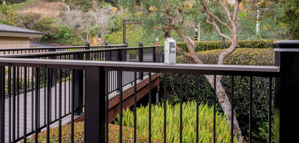 A box of water sites on top of TimberTech’s Drink Rail with black railing and landscaping behind the railing. 