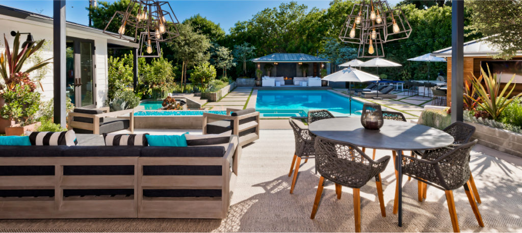 Under an elevated deck includes a couch and dining table with a pool and cabana in the distance.