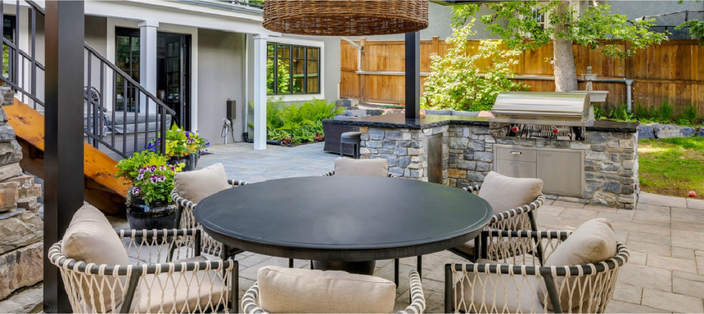 A patio table site under an elevated deck with a rattan lighting fixture above and grill with countertops nearby. 