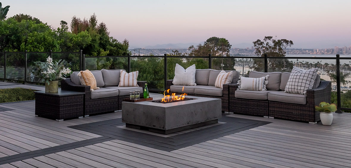 A modern deck fireplace burns in a deck seating area with a bay in the background.