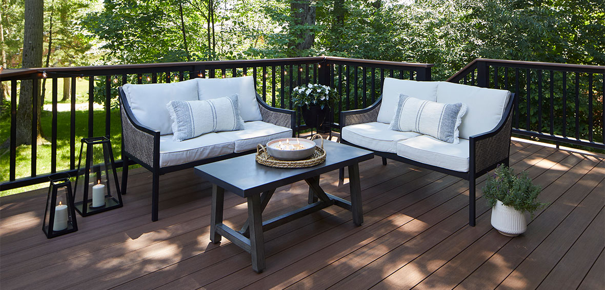Neutral deck furnishings on a dark deck with greenery in the background and potted plants.