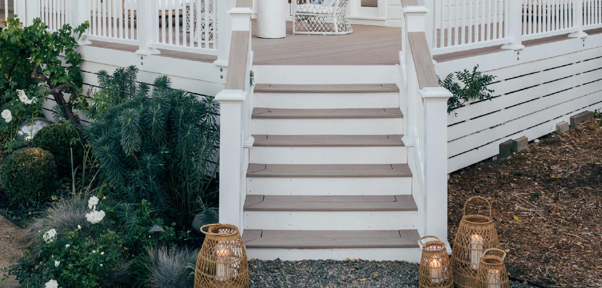 Steps up to a deck showcase wide, flat-top drink rails with landscaping to the side and woven candle lanterns as decor.