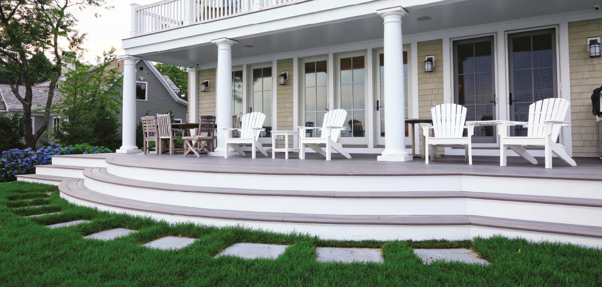 Wide, wraparound steps stretch the length of a back deck with stone steps in the lawn and white chairs on top.