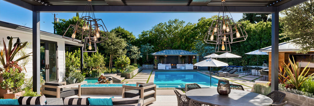 A deck seating and dining arrangement is protected by a pergola next to an inground pool with creative light fixtures overhead.