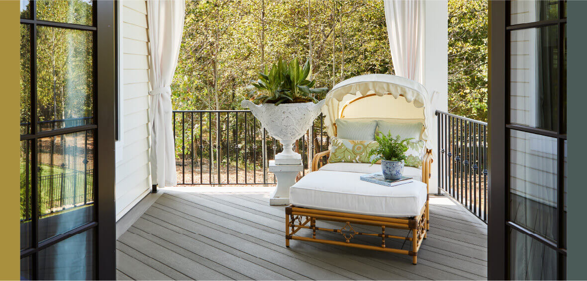 A cushioned chair and ottoman sit on an elevated deck with white curtains tied in the background near the railing.