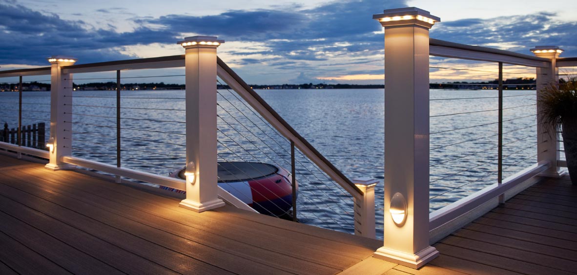 Cable railing infills provide a clear view of a neighboring lake at dusk with soft lights on rail posts and caps.