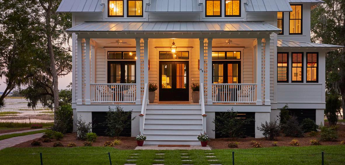 A white, two-story home at dusk has warm lights on and a simple stair design up to a classic-looking front porch.