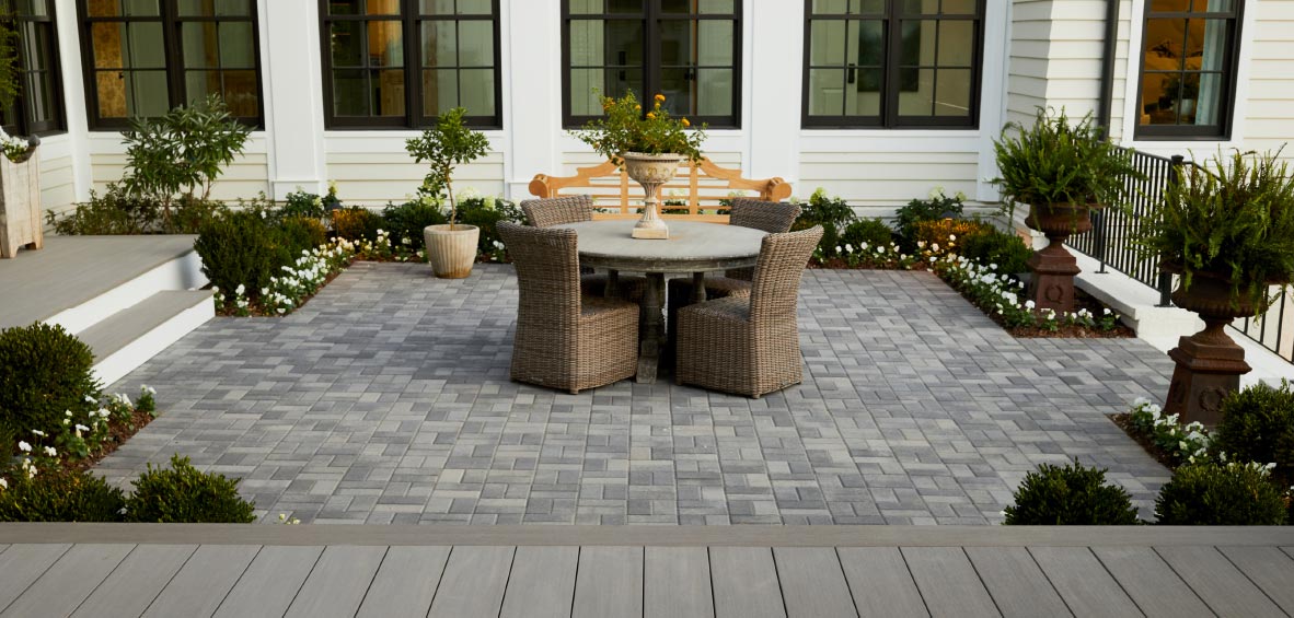 A stone patio shows a dining set in the center with landscaping along each side and steps up to nearby composite decking.