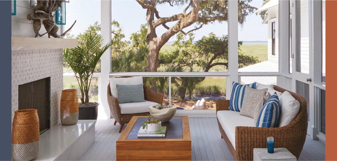 An enclosed sunroom includes a couch, armchair, and coffee table facing a fireplace.
