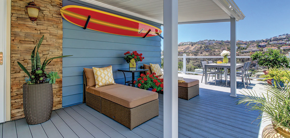 A small deck features two chaise loungers with potted plants of different sizes, tables to elevate decor, and a large surfboard on the wall.