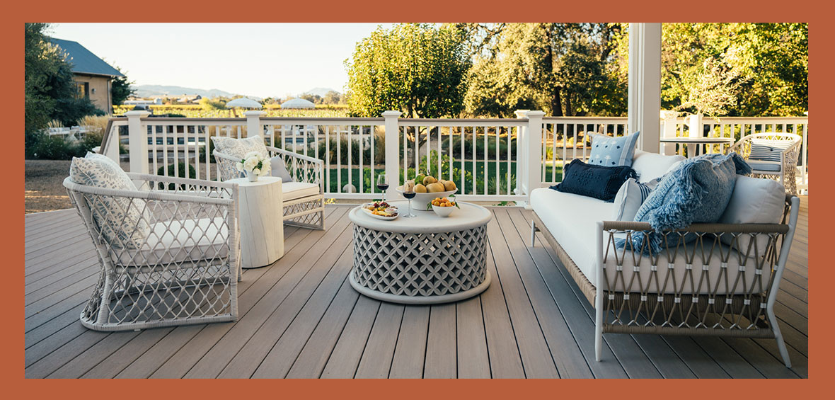 A living room set with a couch, chairs, and table is centered on a backyard deck, with more furniture and trees in the background.