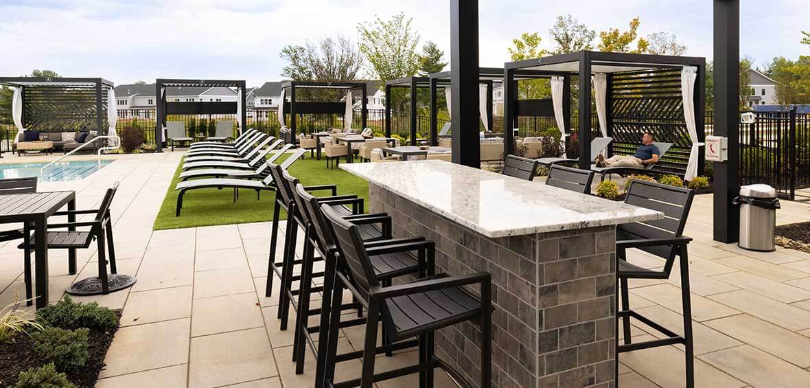 An outdoor space shows a variety of deck furniture around a pool, including lounge chairs and pergolas, with a focus on a bar and seating.