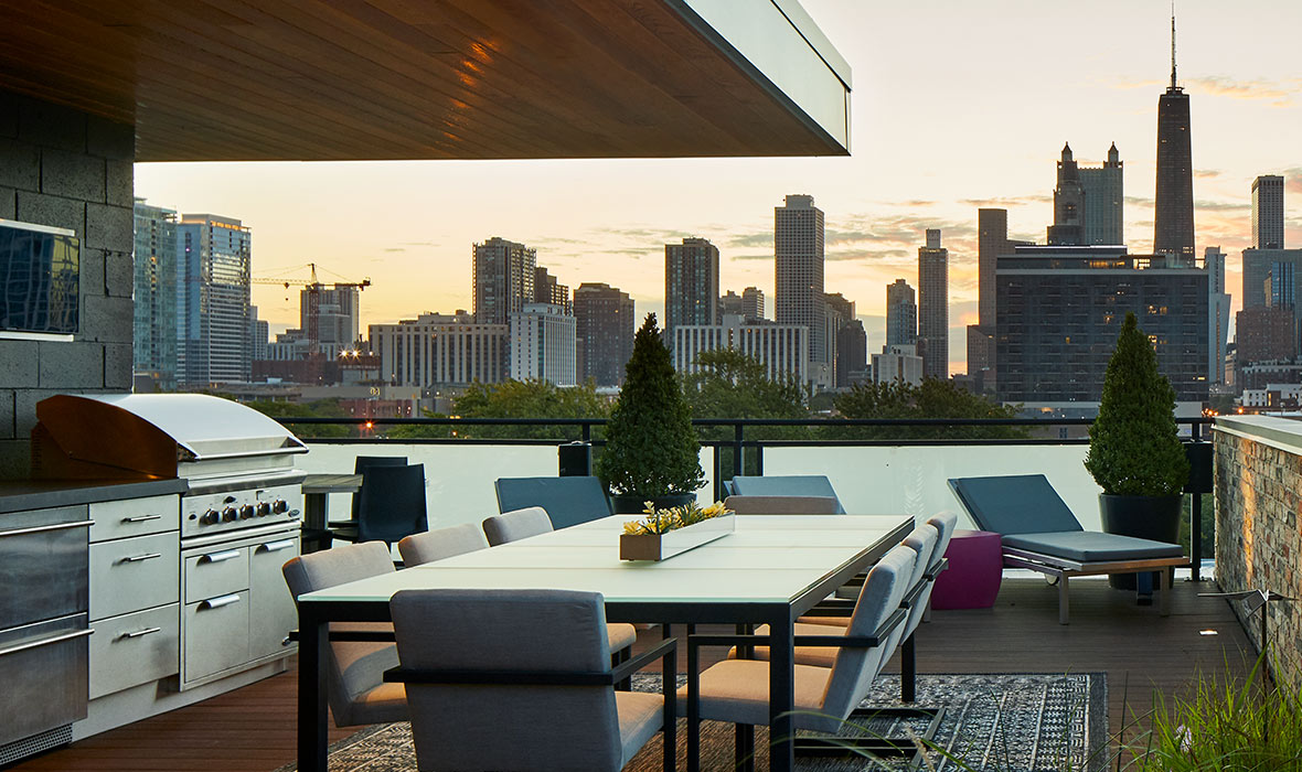 A dining table, bar, barbecue, and lounge chairs are shaded from the sun on a rooftop patio.