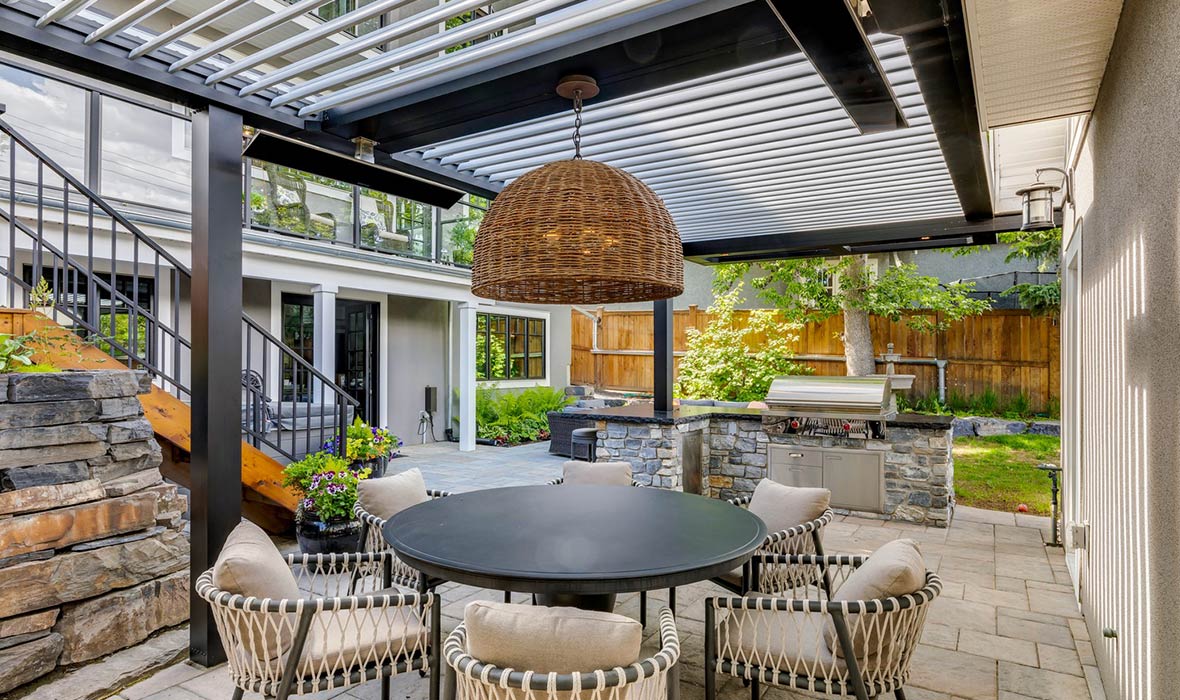 A patio includes a dining area, barbecue, and overhead lighting, all shaded from the sun underneath a pergola.